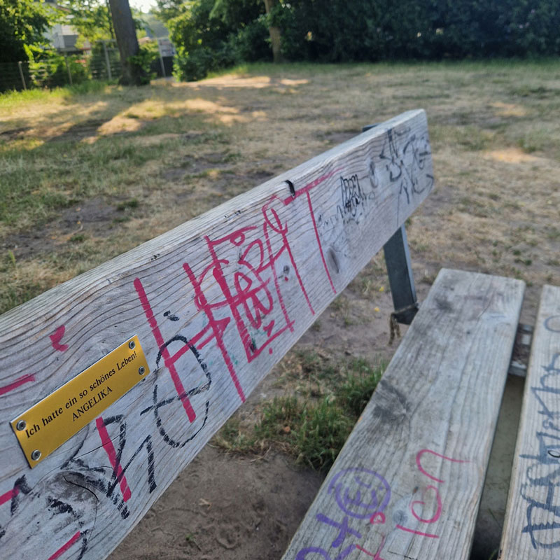 Parkbank mit einem Metallschild auf dem steht: „Ich hatte ein so schönes Leben! – ANGELIKA“